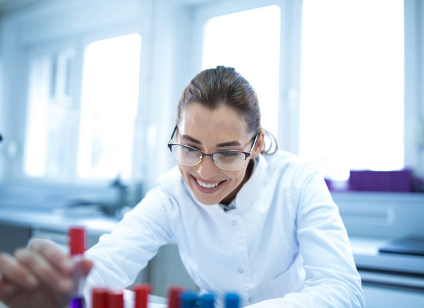 Joven científico técnico de laboratorio trabajando sosteniendo el frasco sonriendo. Mujer satisfecha en el hospital de laboratorio haciendo investigación experimentando.  - Foto, Imagen