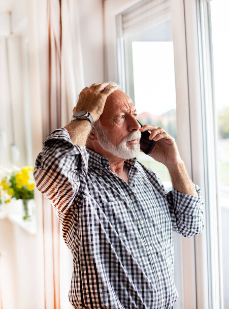 Senior man voor het raam kijkend naar buiten pratend aan de telefoon bezorgd met de hand op het hoofd. - Foto, afbeelding