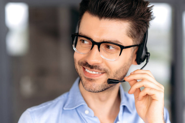 Close up portrait of successful joyful young caucasian operator or call center manager wearing glasses, in formal attire, using headset for video conference or phone call, looking away, smiling - Photo, image