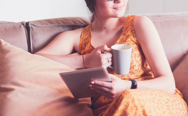 Young woman uses tablet while relaxing at home with coffee on the sofa couch - Фото, изображение