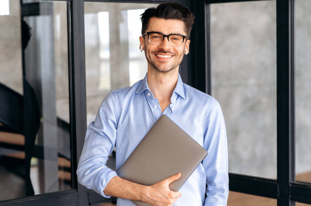портрет привлекательного молодого кавказского бизнесмена или офисного работника, в рубашке и очках, стоящего в современном офисе с ноутбуком, смотрящего в камеру и дружелюбно улыбающегося - Фото, изображение
