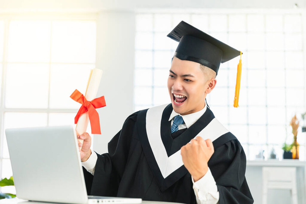 Gelukkige mannelijke afstuderen kijken naar de laptop en het houden van een diploma en het vieren van succes  - Foto, afbeelding