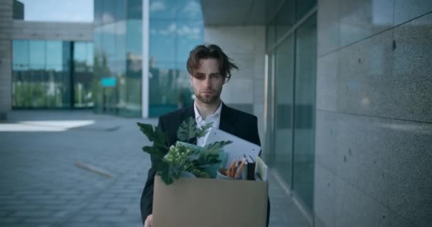 Portrait extérieur de l'employeur masculin licencié déprimé sortant de l'immeuble de bureaux avec une boîte de choses personnelles - Séquence, vidéo