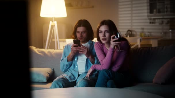 Молодая равнодушная пара проводит вечер дома, женщина смотрит телевизор и пьет вино, мужчина болтает на смартфоне - Кадры, видео