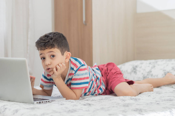 Nuori poikalapsi makaa sängyllä kannettavan tietokoneen avulla. Online-oppitunti videolla. Kaukainen etäopetus. Uusi normaali koulututkimus. Lapset koulussa - Valokuva, kuva