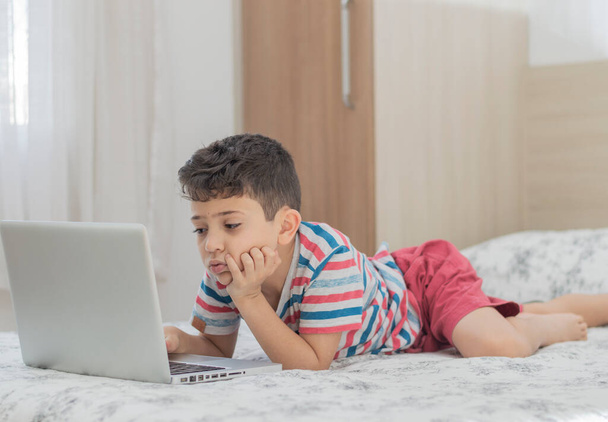Νεαρό παιδί ξαπλωμένο στο κρεβάτι χρησιμοποιώντας ένα φορητό υπολογιστή. Μάθημα online σε βίντεο. Μακρινή εξ αποστάσεως εκπαίδευση. Νέα κανονική σχολική μελέτη. Τα παιδιά στο σχολείο - Φωτογραφία, εικόνα