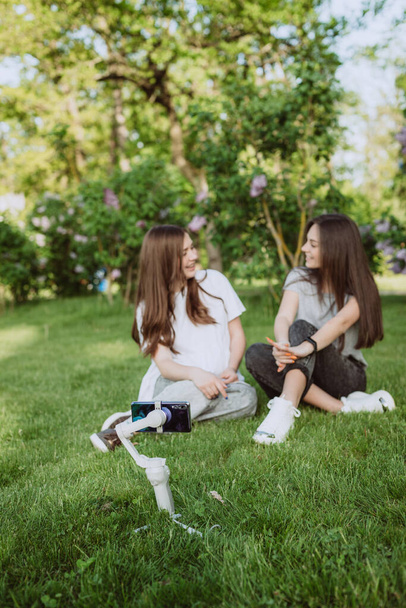 Gülümseyen genç bayan etkili blogcular dışarıda güneşli yeşil bir parkta cep telefonlarıyla bir dengeleyiciye video kaydediyorlar. Blog konsepti. Yumuşak seçmeli odak, odağını kır. - Fotoğraf, Görsel