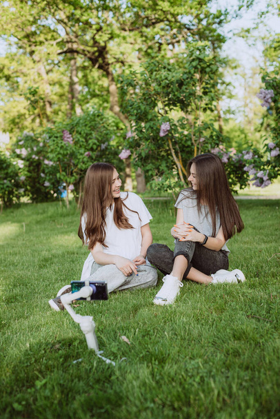 Gülümseyen genç kadın etkili blogcular dışarıda güneşli yeşil bir parkta cep telefonlarıyla bir dengeleyiciye video kaydediyorlar. Blog konsepti. Yumuşak seçici odak. - Fotoğraf, Görsel
