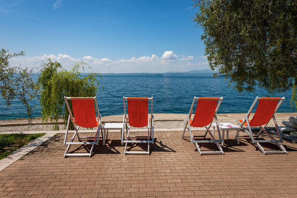 Vier leere Liegestühle an einem wunderschönen Strand am Gardasee (Lago di Garda) zwischen dem kleinen Dorf Bardolino und der Stadt Garda. Provinz Verona, Venetien, Italien, Europa. Lombardische Küste am Horizont. - Foto, Bild