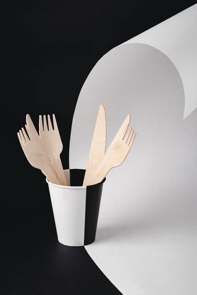 黒と白の背景に紙コップで木製のフォークやナイフ。自然素材の使い捨て食器を環境に配慮。生態学的概念. - 写真・画像