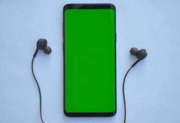 Colpo ad alto angolo di smartphone con schermo verde e auricolare di colore nero su sfondo bianco  - Foto, immagini