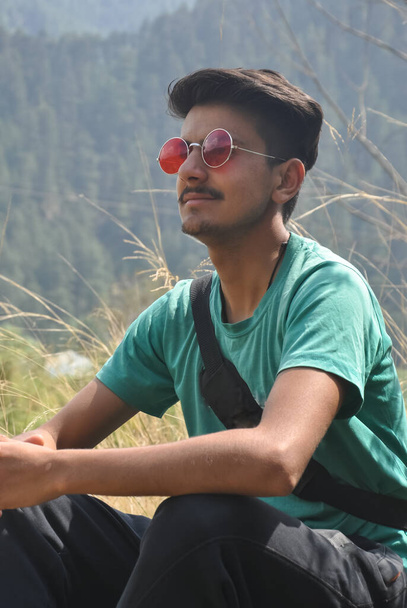 Foto di un bel ragazzo indiano che indossa occhiali da sole e sorride mentre è seduto all'aperto nella natura - Foto, immagini