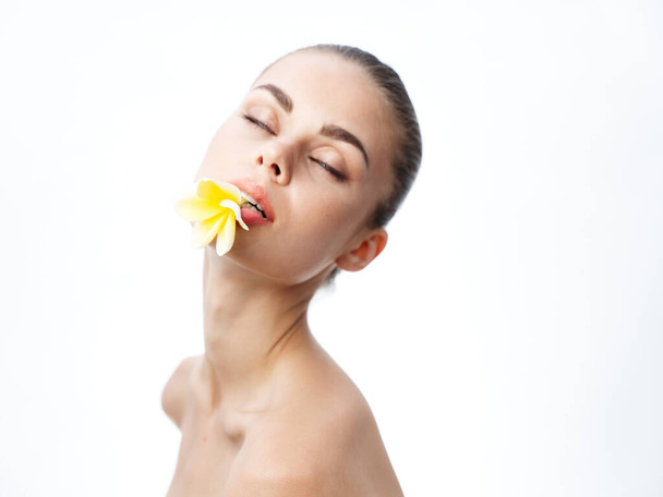 atractiva mujer con hombros desnudos sosteniendo flor amarilla en sus labios y ojos cerrados - Foto, imagen