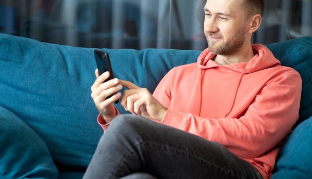 Een man gebruikt een smartphone op zijn thuisbank terwijl hij thuis ontspant. Toepassingen, gesprekken, internet concept. - Foto, afbeelding