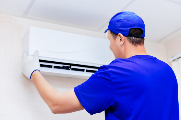 A neat technician in a blue baseball cap and overalls fixes the new air conditioner to the wall in the room.  - Fotó, kép