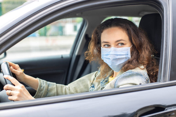 Young woman in a mask sitting in a car, protective mask against coronavirus, driver on a city street during a coronavirus outbreak. - Fotó, kép