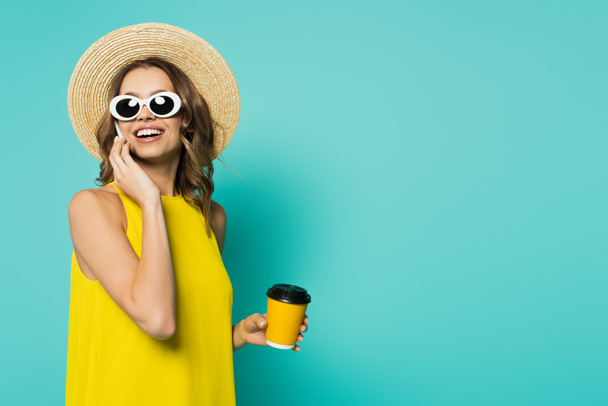 Güneş gözlüklü neşeli kadın elinde kahve bardağıyla mavi arka planda cep telefonuyla konuşuyor.  - Fotoğraf, Görsel
