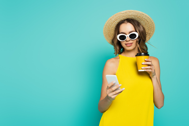 Усміхнена жінка в сонцезахисних окулярах тримає каву, щоб піти і використовувати смартфон ізольовано на синьому
  - Фото, зображення