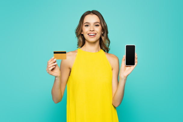 Fröhliche Frau hält Kreditkarte und Handy isoliert auf blauem Grund  - Foto, Bild