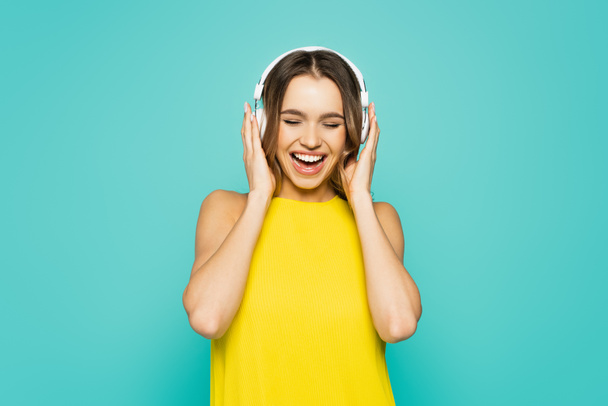 Positive woman in yellow dress listening music in headphones isolated on blue  - Fotó, kép