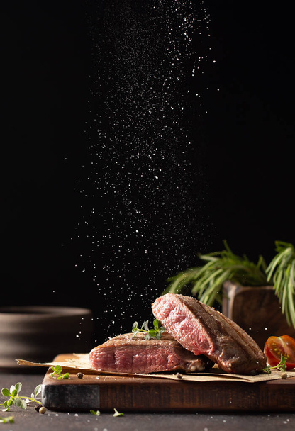 Cut beef steak on a wooden board with fresh thyme,rosemary,cherry tomatoes,peppercorns and falling sea salt.Close up of meat dish in a freeze motion. - Foto, immagini