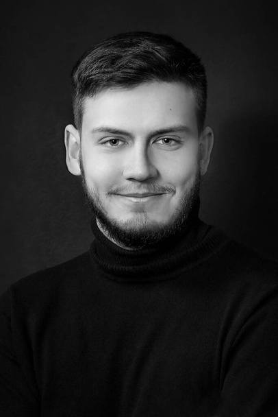 black and white studio portrait of a blue-eyed smiling young handsome man with a beard in black clothing on a black background;  look directly into the camera lens; vertical - Photo, image