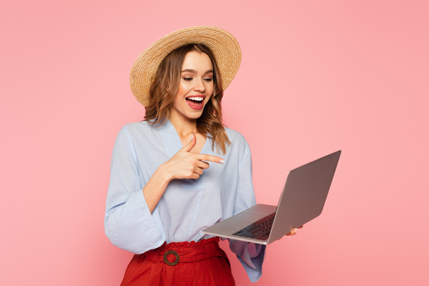 Mulher feliz em chapéu de palha apontando com o dedo para laptop isolado em rosa  - Foto, Imagem