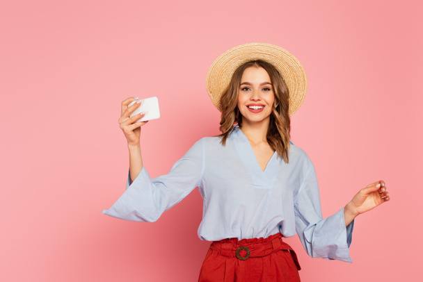 Pretty woman in sun hat holding smartphone and looking at camera on pink background  - Foto, immagini