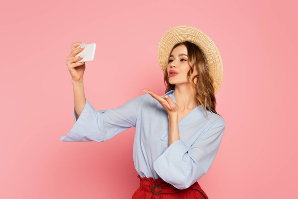 Pretty woman in sun hat blowing air kiss while taking selfie on smartphone isolated on pink  - Фото, изображение