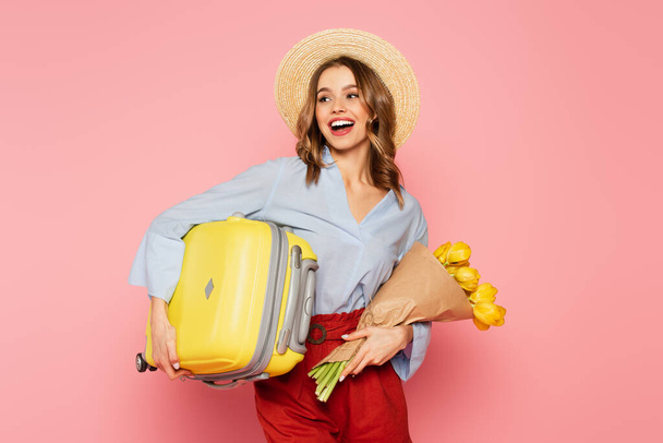 Positive woman in straw hat holding tulips and suitcase isolated on pink  - Foto, Bild