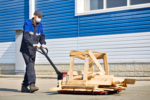 Mężczyzna w ubraniu roboczym i masce medycznej przewozi ładunek na wózku na terytorium bazy przemysłowej w letni dzień. - Zdjęcie, obraz