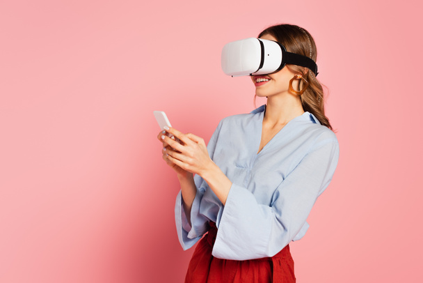 Cheerful woman in vr headset holding smartphone on pink background  - Foto, afbeelding
