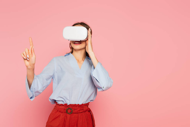 Smiling woman in vr headset pointing with finger isolated on pink  - Φωτογραφία, εικόνα