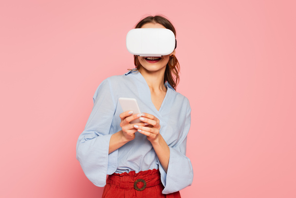 Cheerful woman in vr headset using mobile phone isolated on pink  - Foto, imagen