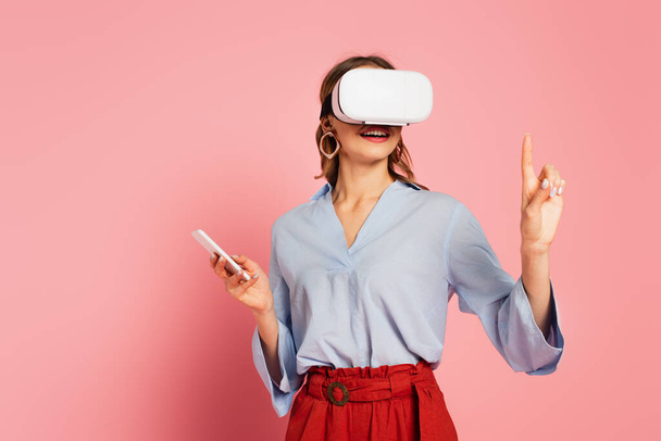 Smiling woman in vr headset pointing with finger on pink background  - Foto, imagen