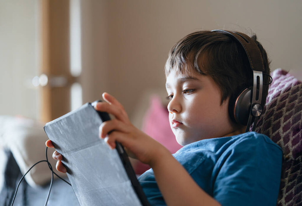 Dziecko leżące na kanapie, noszące słuchawki słuchające muzyki, zbliżenie twarzy Dziecko siedzące na kanapie oglądające kreskówki na tablecie, Młody chłopiec grający w grę na dotyku lub relaksujący się samodzielnie w salonie - Zdjęcie, obraz