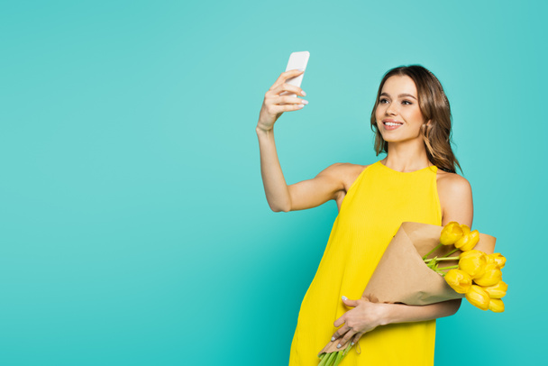 Happy woman with yellow tulips talking selfie on smartphone isolated on blue  - Photo, image