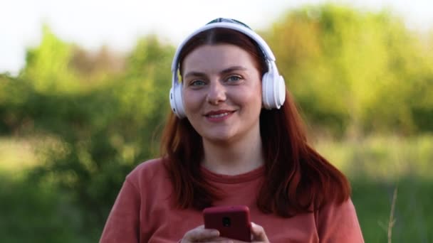 Caucasico millenario felice donna sorridente viso utilizzando smartphone e ascoltare musica da cuffie fuori sulla strada all'aperto - Filmati, video