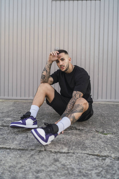 A young man with tattoos in cool casual style sitting on the ground - Фото, зображення