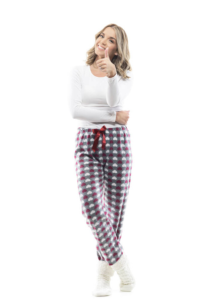Fröhliche junge Frau zu Hause im kuscheligen Pyjama, die den Daumen in die Kamera zeigt. Volle Körperlänge isoliert auf weißem Hintergrund. - Foto, Bild