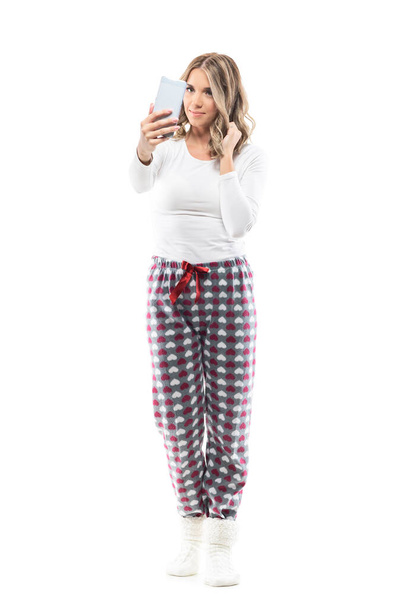 Linda mujer joven en pijama tomando selfie con el teléfono celular tocando el pelo. Longitud total del cuerpo aislado sobre fondo blanco. - Foto, imagen