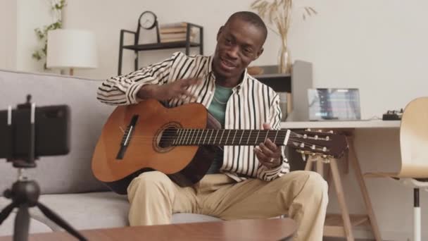 Střední záběr talentovaného afrického mužského hudebníka sedícího na pohovce ve studiu zvukového designu a střílejícího na akustickou kytaru na kameře smartphonu - Záběry, video
