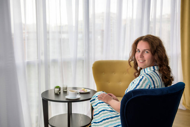 Νεαρή γυναίκα στο σπίτι κάθεται σε μοντέρνα καρέκλα μπροστά από το παράθυρο χαλαρώνοντας στο σαλόνι της και πίνοντας καφέ ή τσάι με γλυκά ή έρημο. Χαλαρώστε πριν από τη νέα εργάσιμη ημέρα. - Φωτογραφία, εικόνα