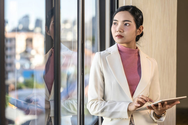 Ázsiai üzletasszony alkalmi öltöny álló és tartó technológia tabletta mellett az ablak üveg, üzleti és életmód koncepció - Fotó, kép