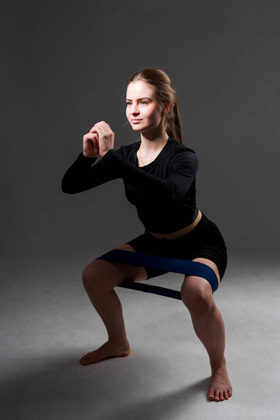 Beautiful woman in a black sweater and sports shorts doing squats using an elastic band expander - Фото, изображение