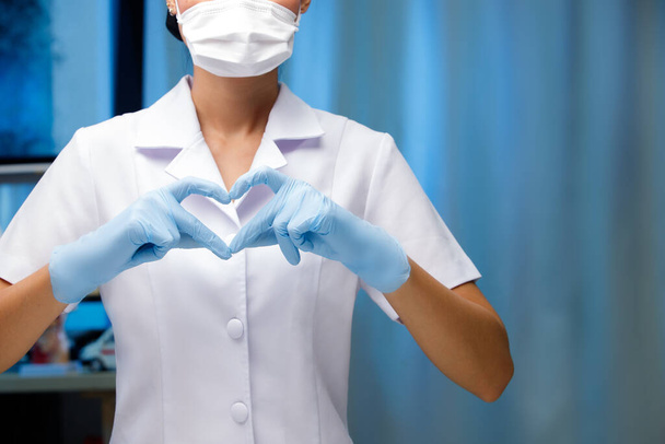 Infirmière Médecin en masques médicaux et gants en caoutchouc montrant la forme du cœur avec les mains. Contexte de l'hôpital de ton bleu dans le concept cardiovasculaire - Photo, image