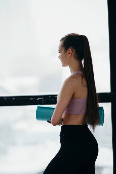 Photo sideways. A girl in a sports top and leggings stands against the background of a window and holds a yoga mat in her hands - Фото, зображення