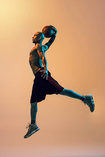 Full length portrait of african-american male basketball player in action in neon light on orange background. - 写真・画像