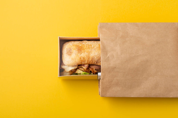 Sandwich en contenedor de comida para llevar o entrega y bolsa de papel sobre fondo amarillo, vista superior - Foto, Imagen