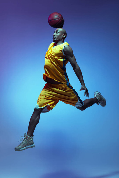 Handsome african-american male basketball player in action and motion in neon light on blue background. Concept of healthy lifestyle, professional sport. - 写真・画像